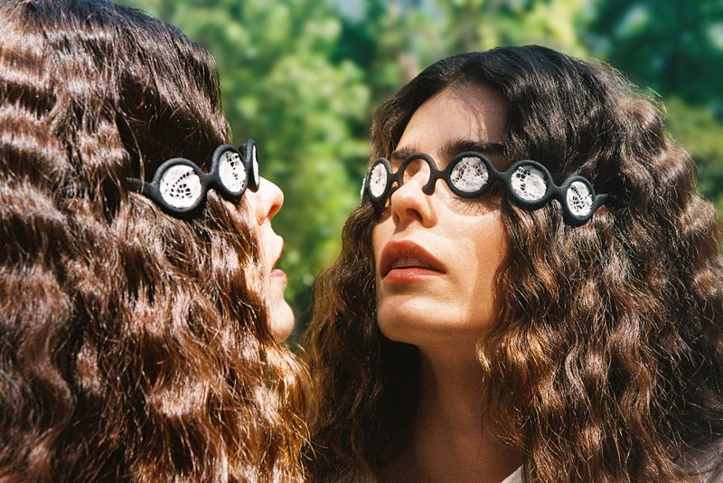 Two women with long crimped dark hair wear avant-garde circular black sunglasses filled with white lace in front of outdoor greenery.