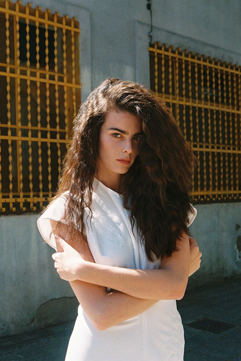 Woman with voluminous wavy brunette hair wearing a white dress and crossing her arms in front of yellow window grates.