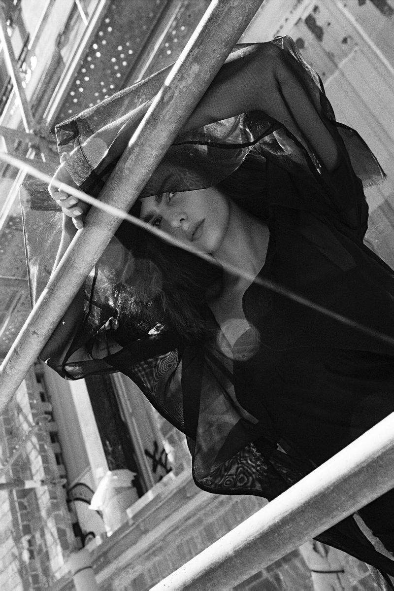 Black and white portrait of a woman posing through scaffolding pipes wearing sheer draped black fabric