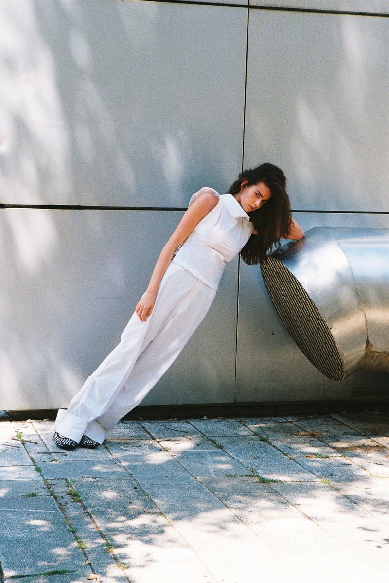 Fashion model in a white sleeveless top and wide trousers leaning at an angle against a large metal duct.
