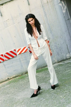 Woman with long crimped hair in an open white tailored blazer and matching wide-leg trousers against a concrete wall.