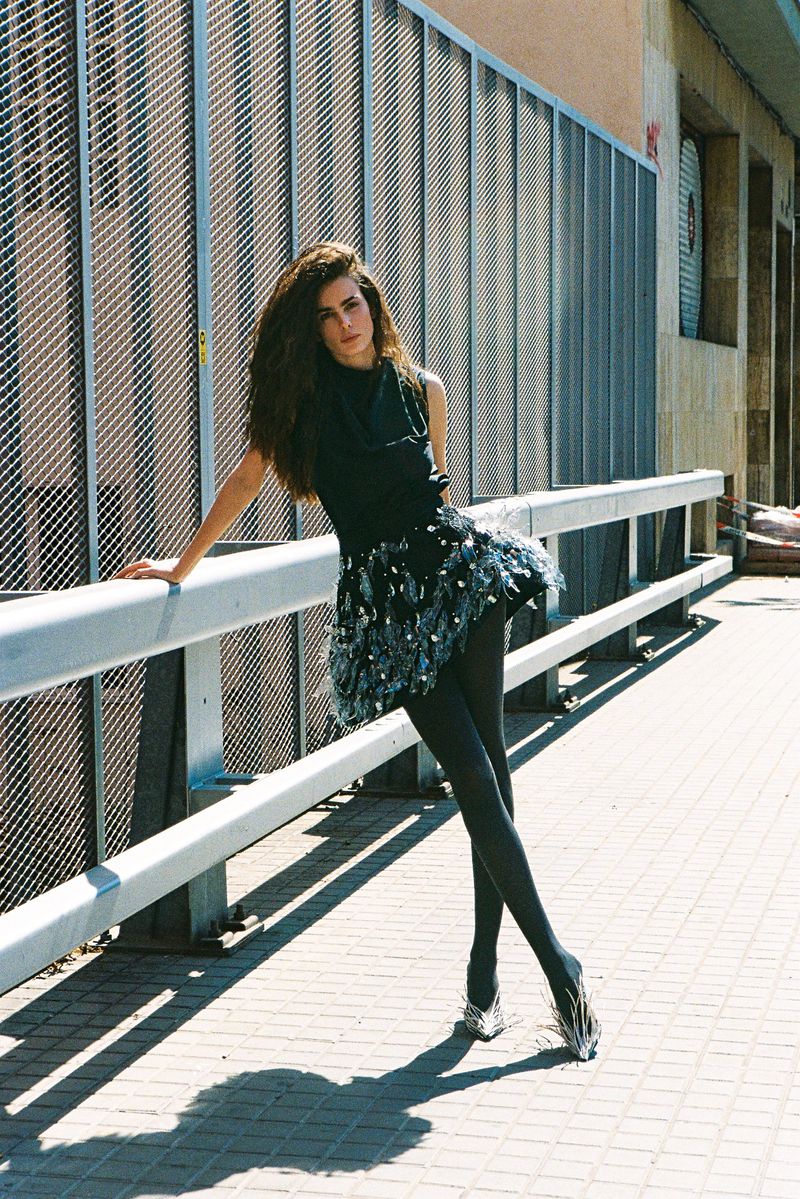 Woman in a black dress with a feathered skirt leaning against a metal railing on an outdoor walkway