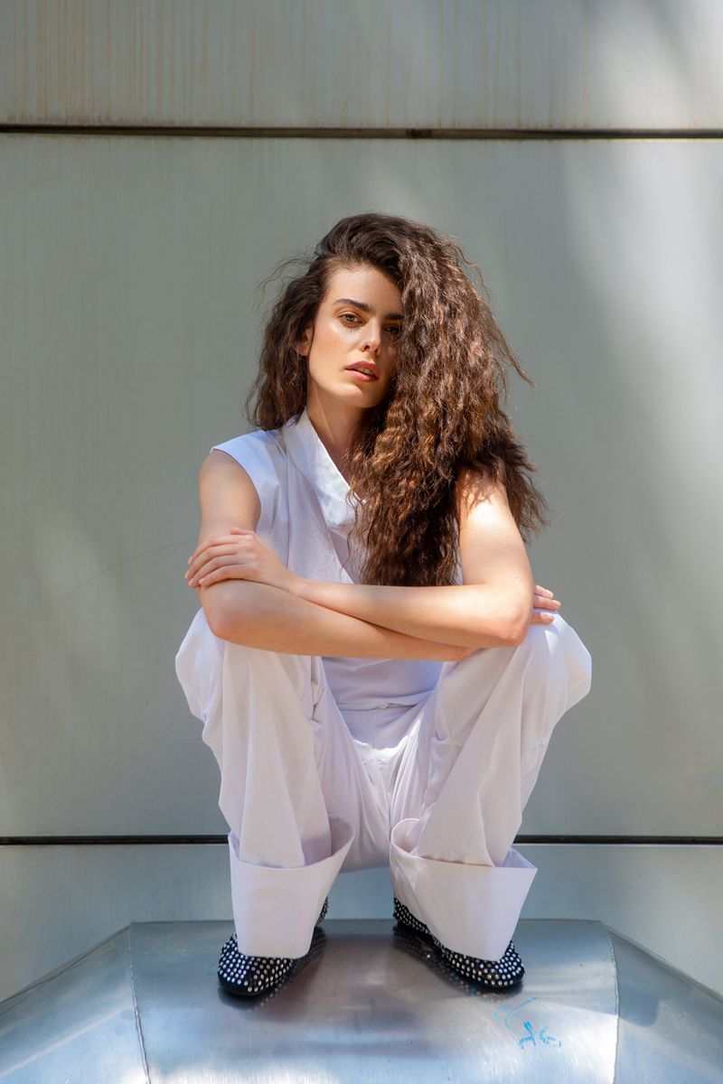 Woman with voluminous curly hair crouching in an all-white sleeveless outfit and wide-cuff trousers against a panelled wall.
