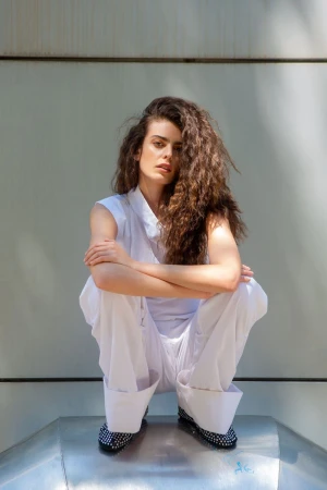 Woman with voluminous curly hair crouching in an all-white sleeveless outfit and wide-cuff trousers against a panelled wall.