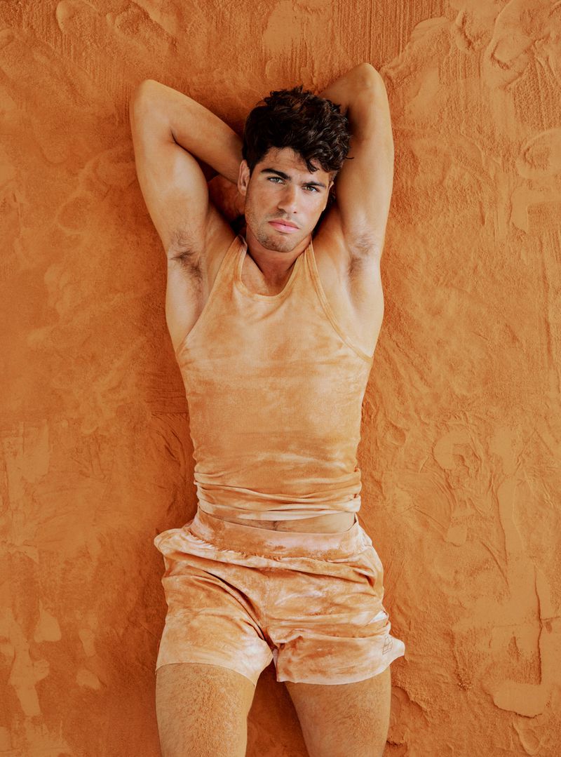 Carlos Alcaraz lying on a textured orange clay surface wearing a matching sleeveless tank top and shorts with hands behind his head.