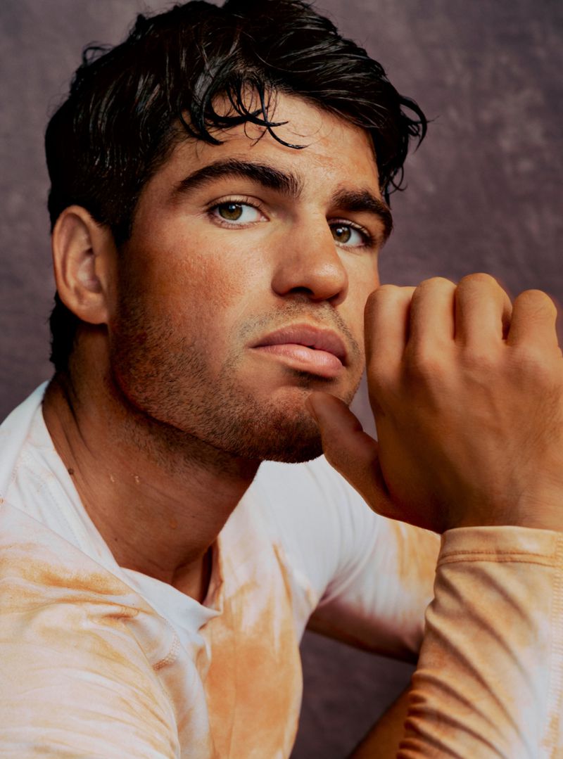 Close-up portrait of Carlos Alcaraz with hand raised near chin wearing a peach and white top against a textured studio backdrop.