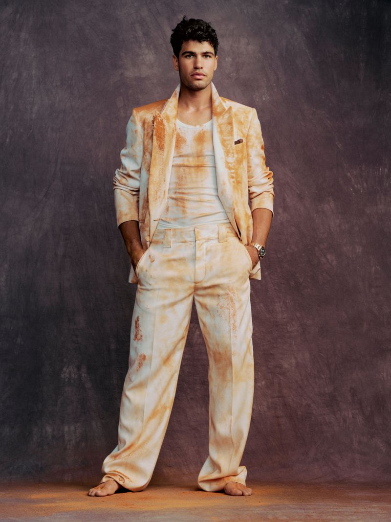 Tennis player Carlos Alcaraz barefoot in a clay-stained beige tailored suit standing against a textured studio backdrop.
