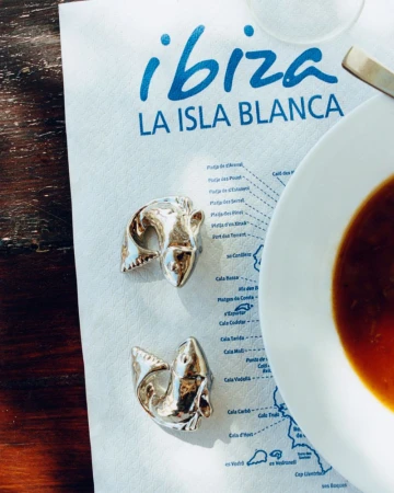 Two metallic silver fish-shaped jewelry pieces resting on an Ibiza paper map next to a bowl of soup on a wooden table.