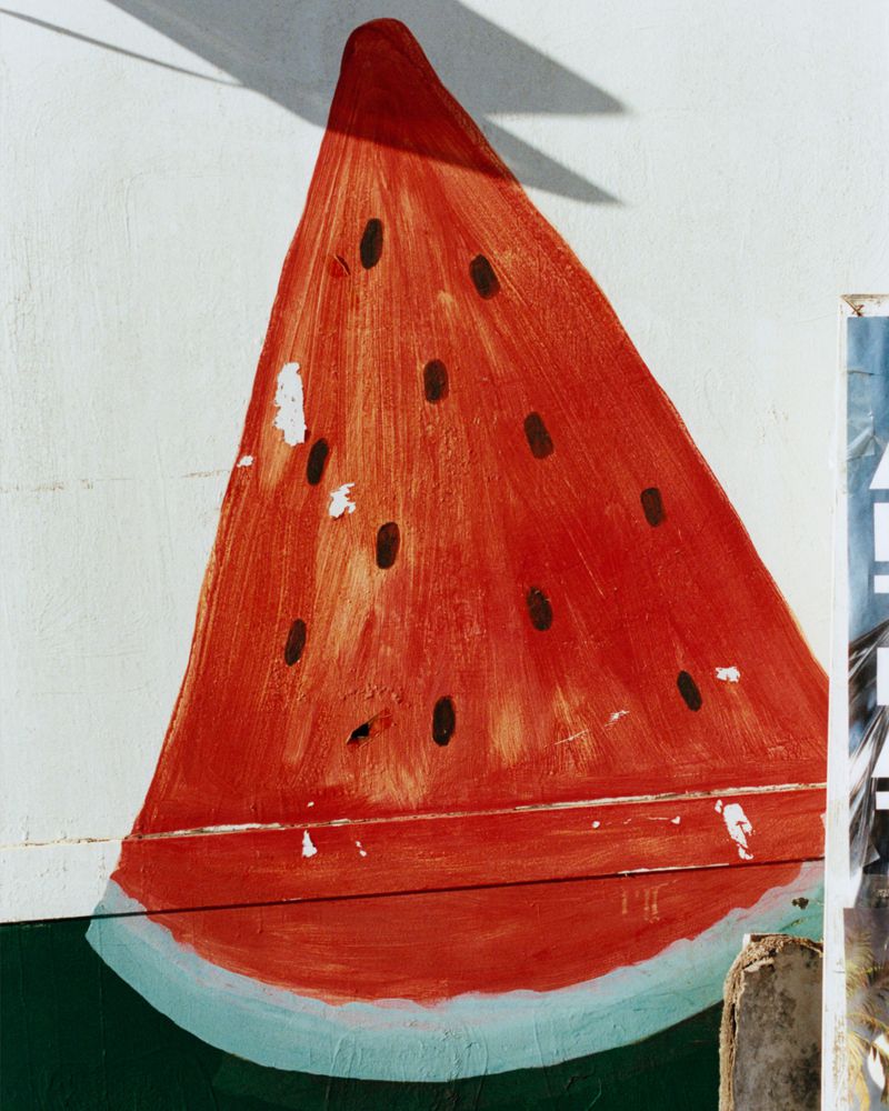 A painted slice of red watermelon with black seeds on a sunlit textured white wall with a sharp cast shadow.