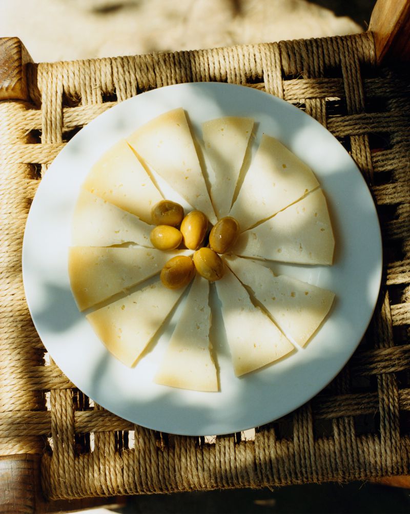 Slices of cheese arranged in a radial circle with olives in the center on a white plate resting on a woven rope chair.