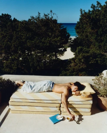 Shirtless man resting on yellow-and-white striped cushions on a sunny terrace overlooking coastal trees and the ocean.