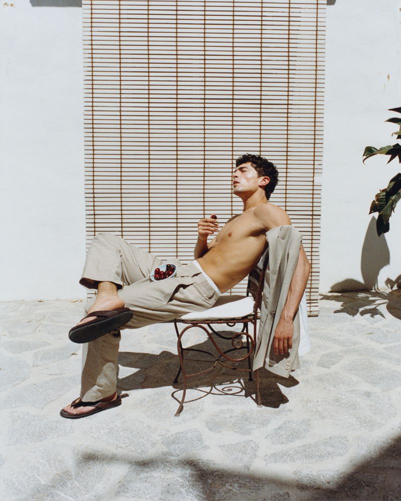 Shirtless man wearing beige trousers and sandals relaxes on an iron chair in bright sunlight for Jacquemus Resorts.