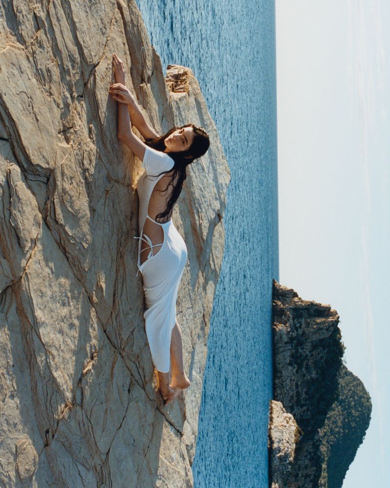 Model Isis Bensalem reclining on a coastal rock formation in a white cutout dress overlooking the sea.