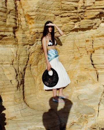 Isis Bensalem shields her eyes in a bikini top and white pleated skirt, holding a round black tote against a textured sandstone cliff.