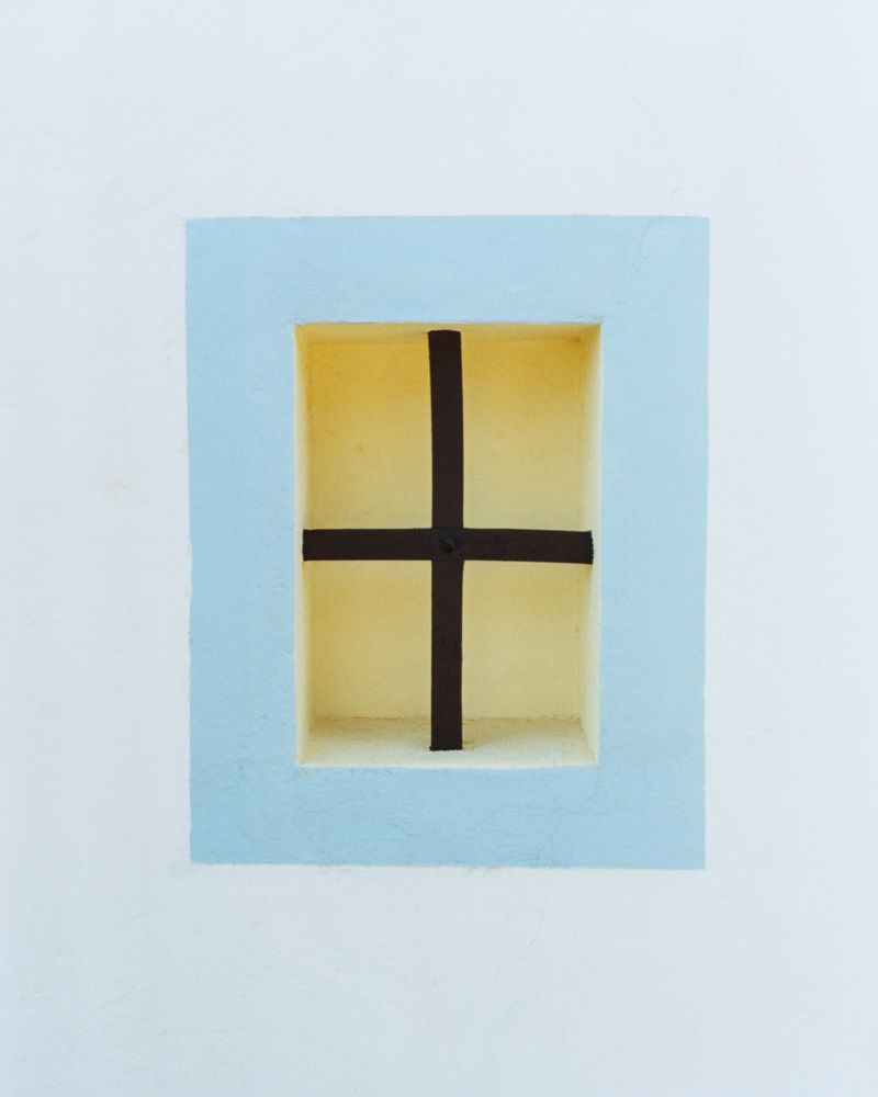 Architectural window detail with a light blue frame, dark cross bar, and pale yellow niche on a white wall.