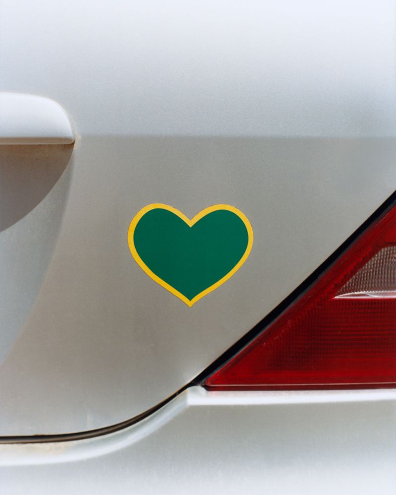 A dark green heart sticker with a yellow border affixed to the white exterior panel of a car next to a red taillight.