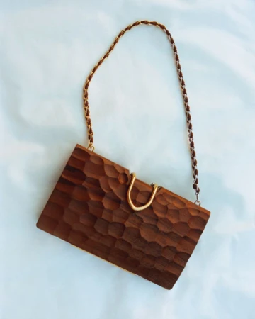 Carved wooden rectangular clutch handbag with a gold-tone clasp and braided chain strap against a pale blue background.