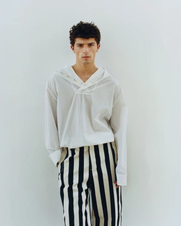 Model Isis Bensalem wearing a loose white sailor shirt and vertical black and white striped trousers against a plain white studio wall.