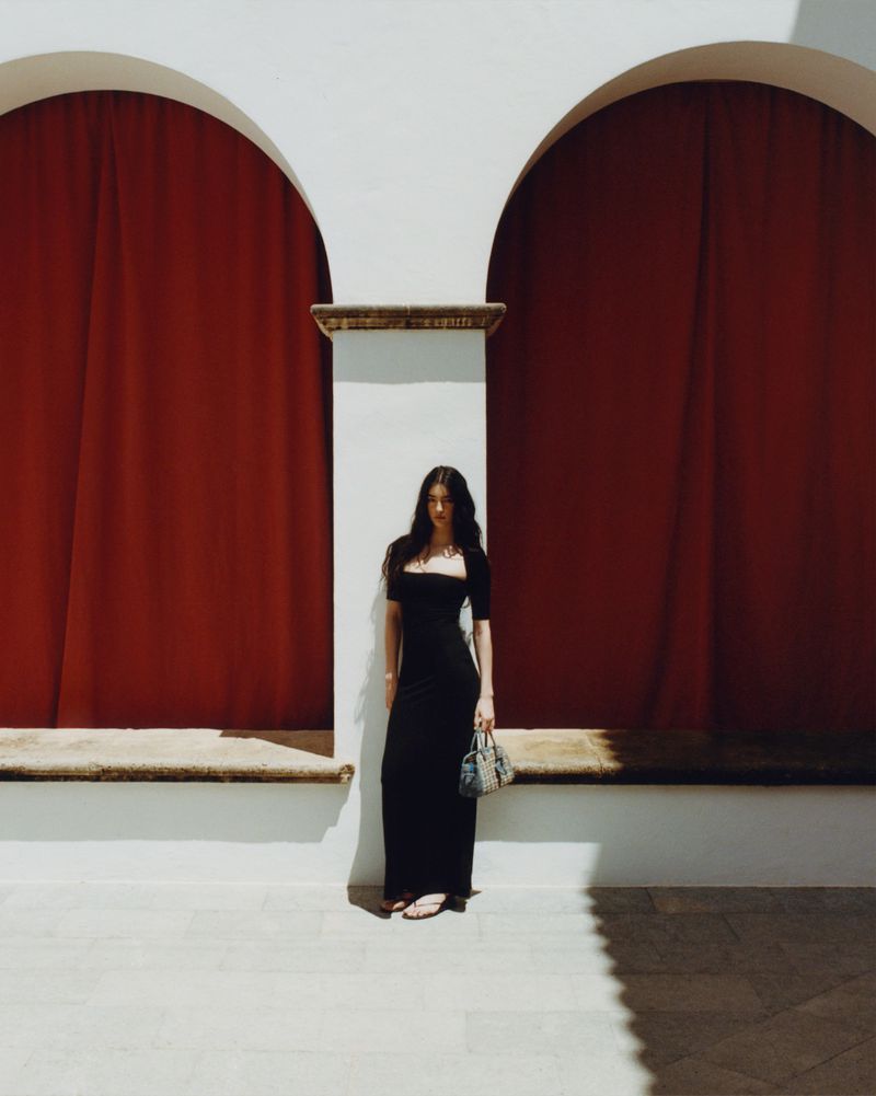 Model Isis Bensalem wearing a black maxi dress and holding a handbag, standing between two white arches with red curtains.