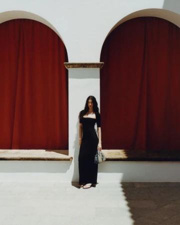 Model Isis Bensalem wearing a black maxi dress and holding a handbag, standing between two white arches with red curtains.