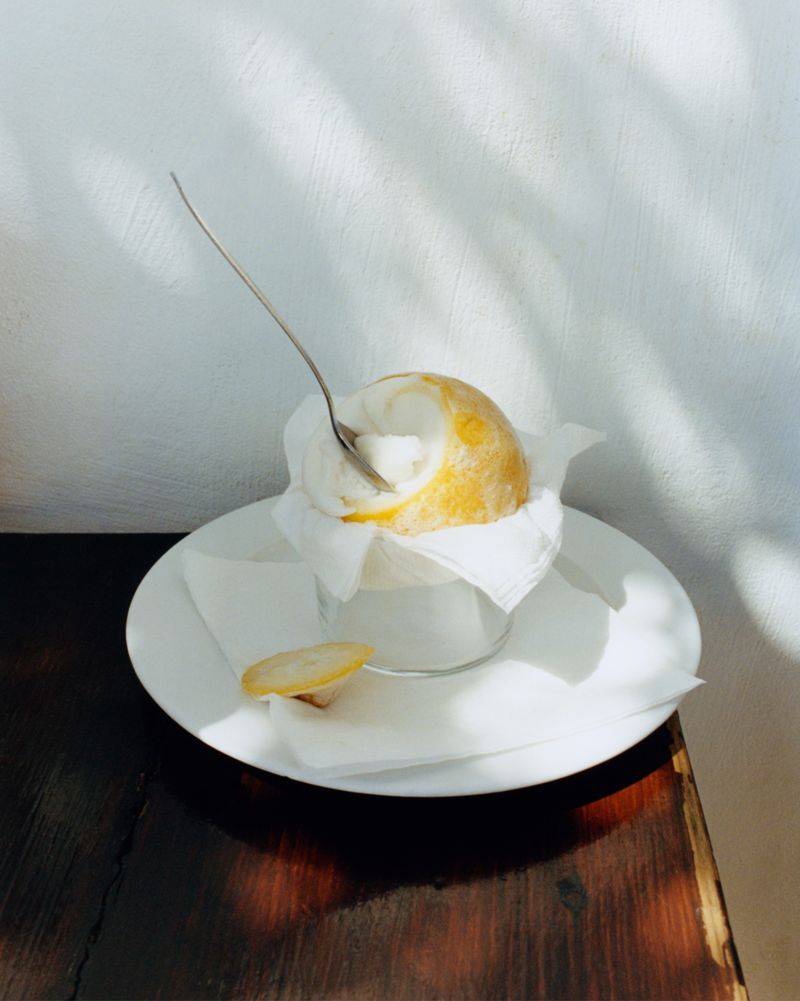 A lemon sorbet dessert served inside a frozen lemon rind with a spoon on a white plate over a dark wooden table.