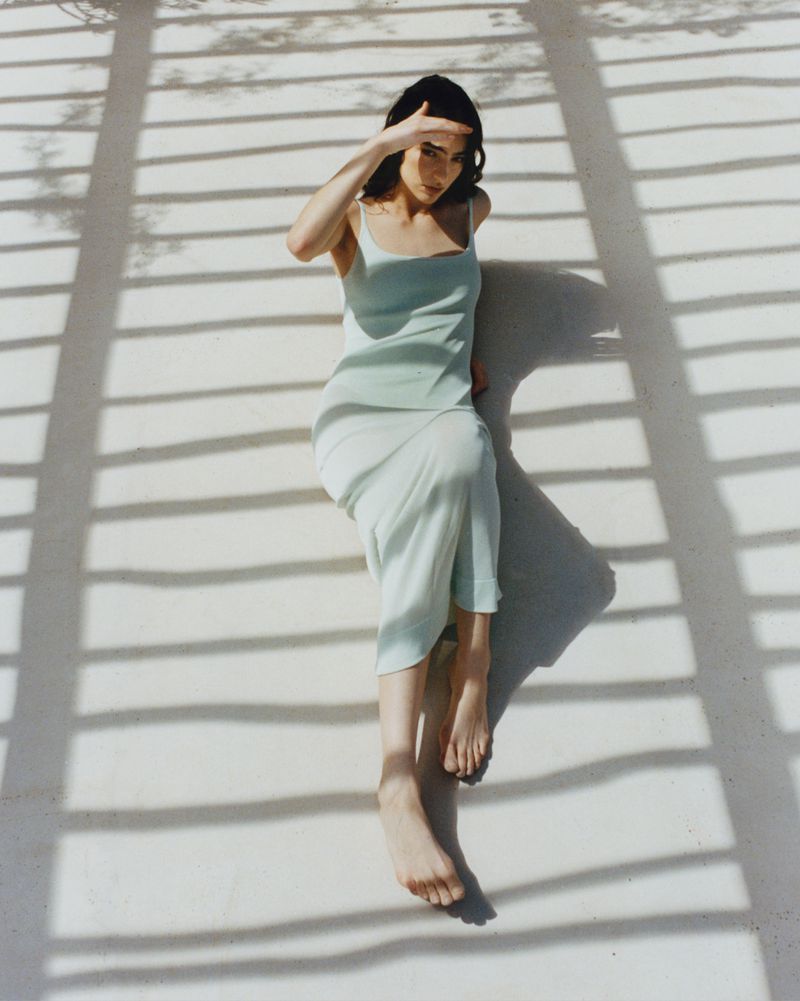 Model Isis Bensalem in a pale mint slip dress reclining outdoors under slatted pergola shadows.
