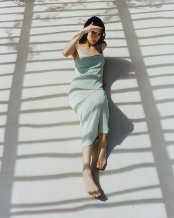 Model Isis Bensalem in a pale mint slip dress reclining outdoors under slatted pergola shadows.