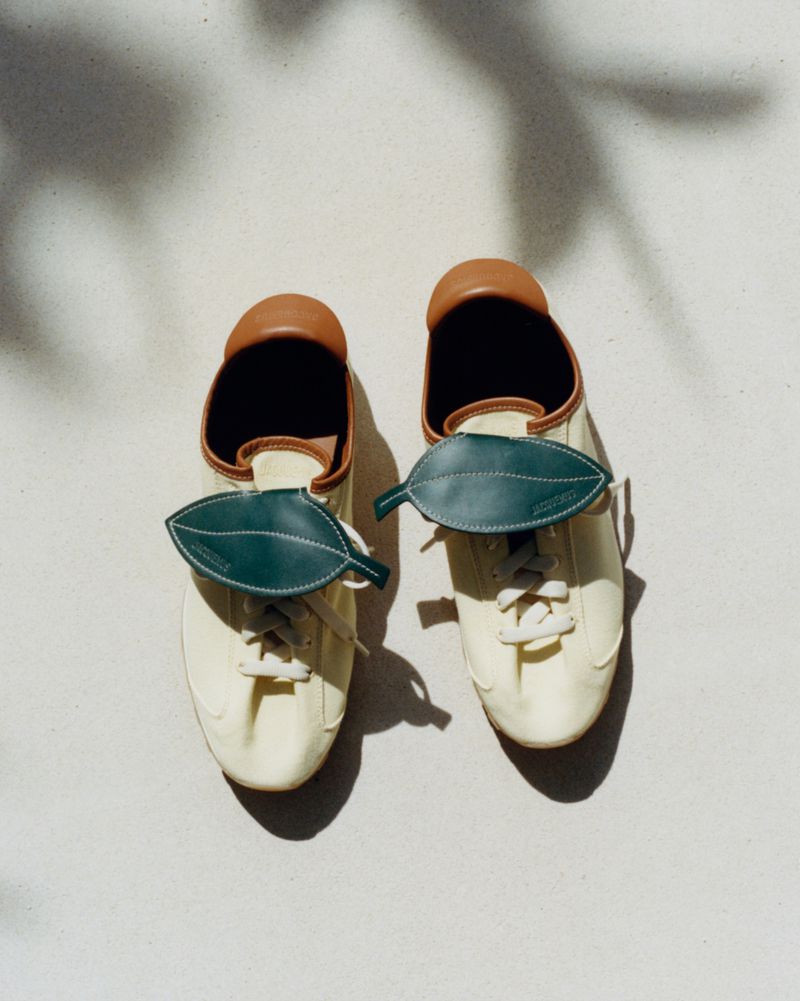 Pair of Jacquemus cream sneakers with dark green leather leaf flaps over the laces resting on a light sunlit surface.