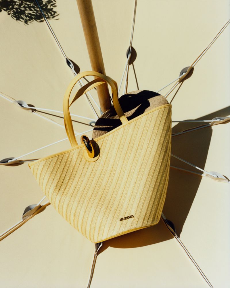 Yellow ribbed Jacquemus tote bag and straw sunhat hanging from a parasol pole under direct sunlight.