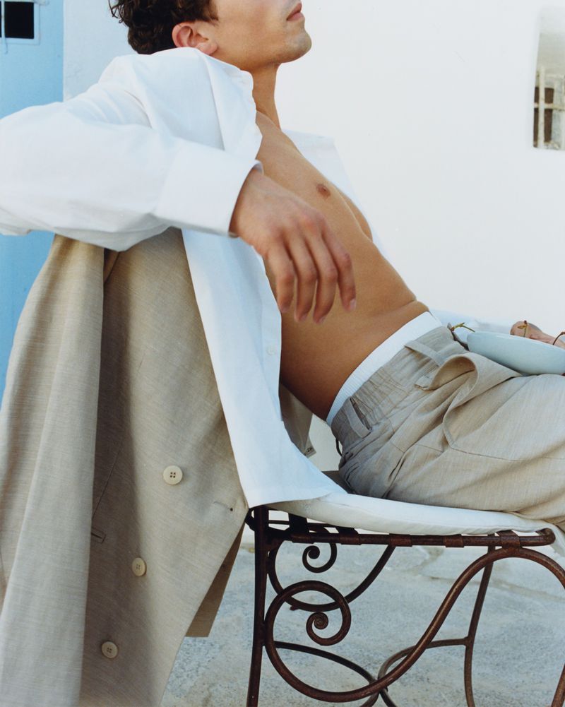 Man in unbuttoned white shirt and beige linen trousers reclining on an ornate wrought-iron chair outdoors against a white wall.
