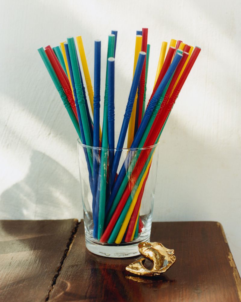 A clear glass filled with colorful flexible plastic straws sits on a dark wooden table beside a small gold ring in bright sunlight.