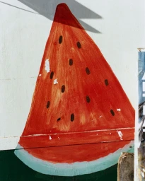 A painted slice of red watermelon with black seeds on a sunlit textured white wall with a sharp cast shadow.