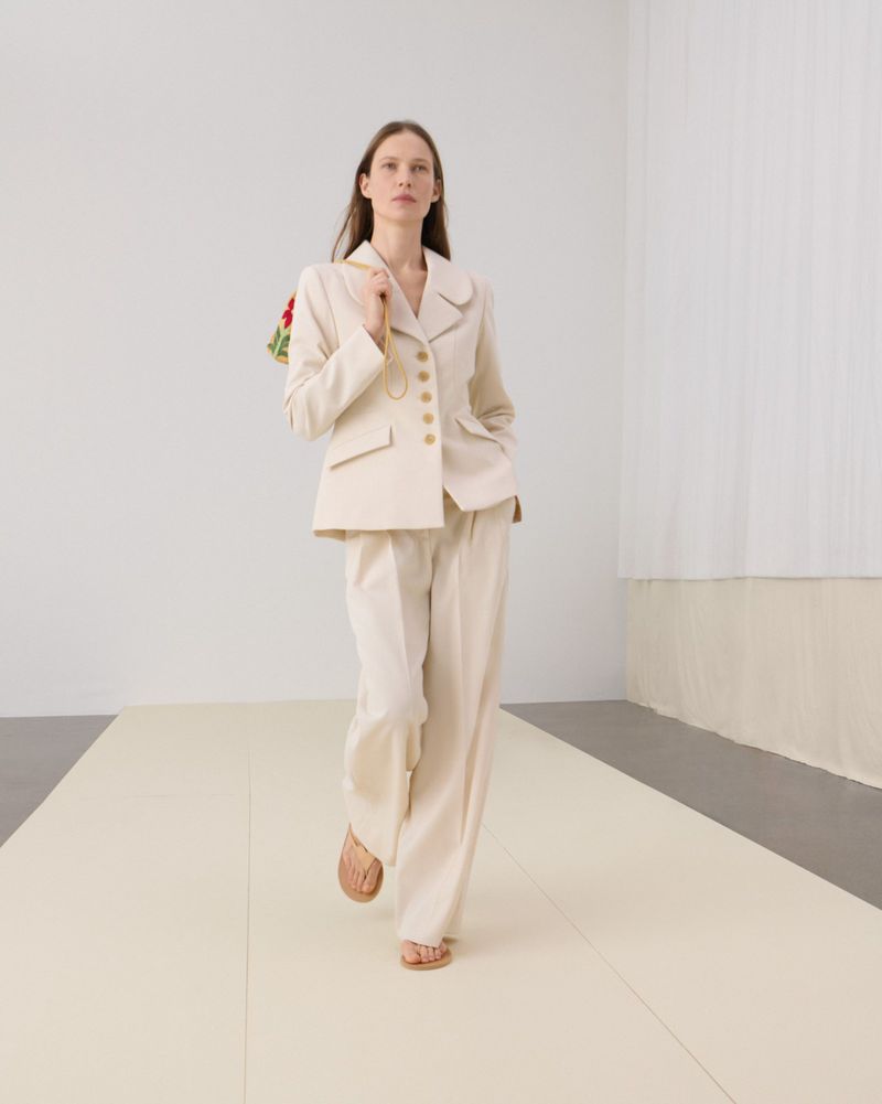 A woman walks in a runway outfit featuring a cream-colored blazer and wide-leg pants, carrying a floral purse in a minimalist setting.