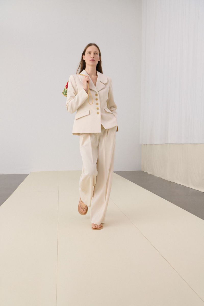 Woman in a beige suit and sandals walking on a beige floor, carrying flowers, in a brightly lit indoor space.