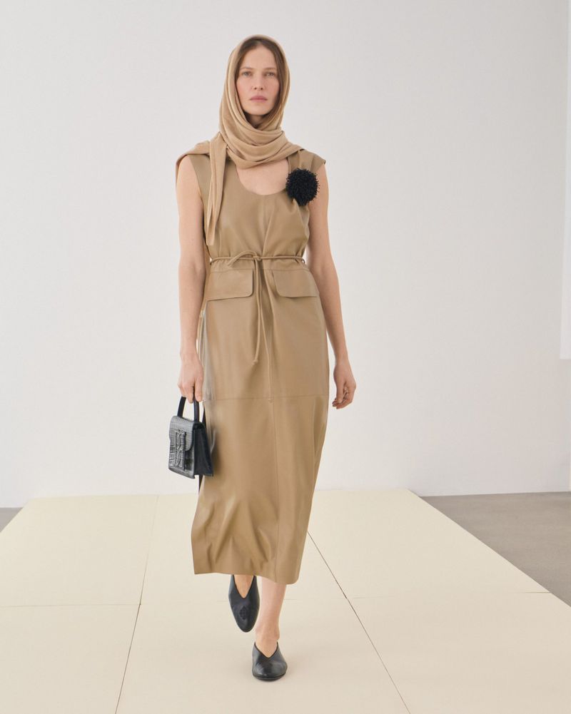 A slender woman in a light brown dress, black flats, and a headscarf walks on a light beige runway, carrying a small black purse in a minimalist fashion image.