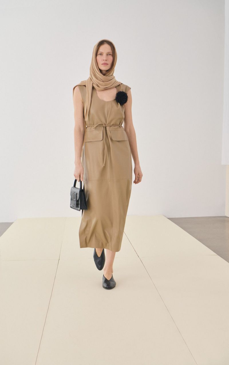 A woman models a sleeveless tan dress with a scarf draped over her head and a black purse in a minimalist, bright studio setting.