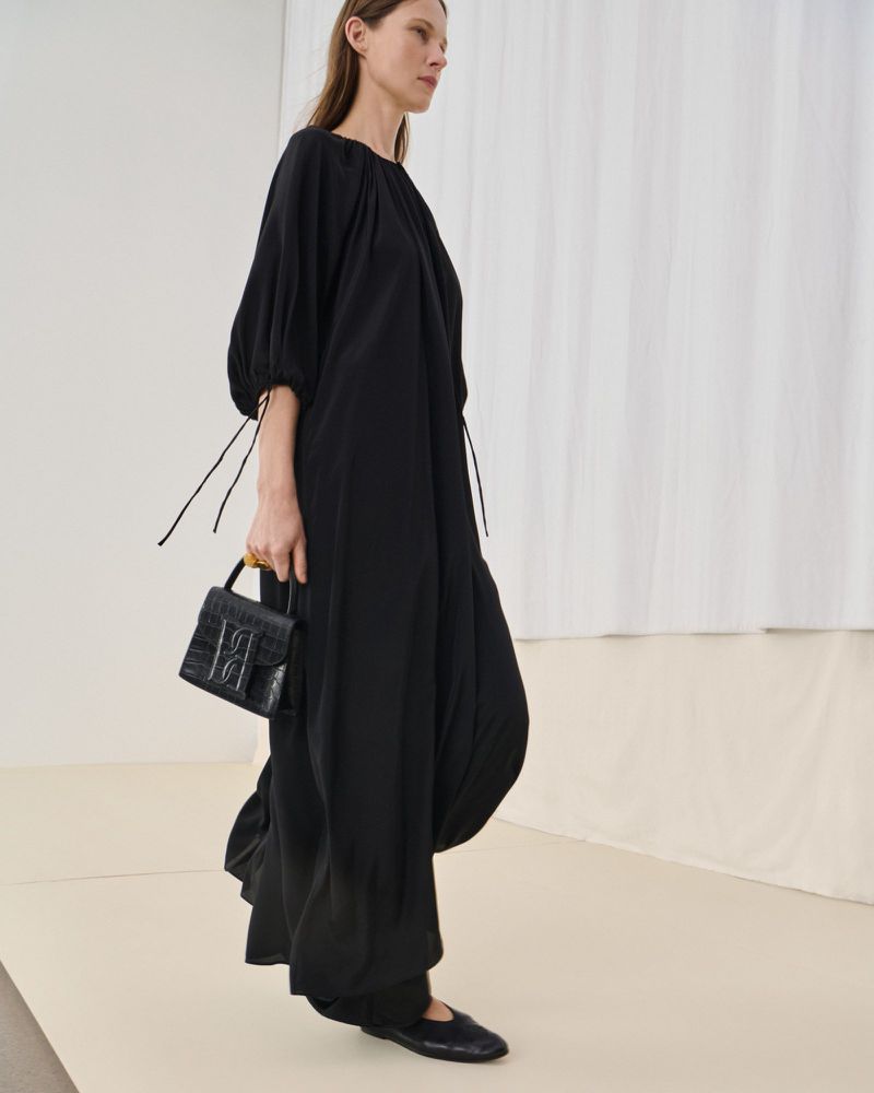A woman in a long black dress carries a black purse in a fashion setting with a white backdrop, showcasing a minimalist aesthetic.