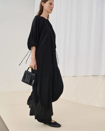 A woman in a long black dress carries a black purse in a fashion setting with a white backdrop, showcasing a minimalist aesthetic.