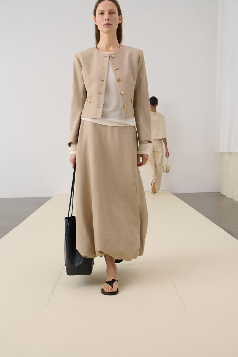 A woman showcases a beige jacket and skirt on a runway, paired with a black tote bag, with another model visible in the background.