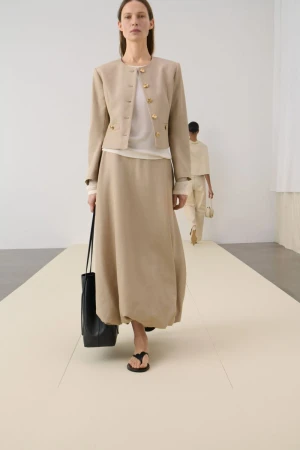 A woman showcases a beige jacket and skirt on a runway, paired with a black tote bag, with another model visible in the background.
