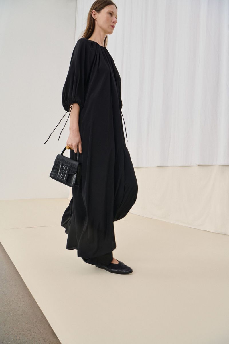 Fashion model in a black dress and matching flats, carrying a leather handbag against a neutral backdrop in a studio environment.