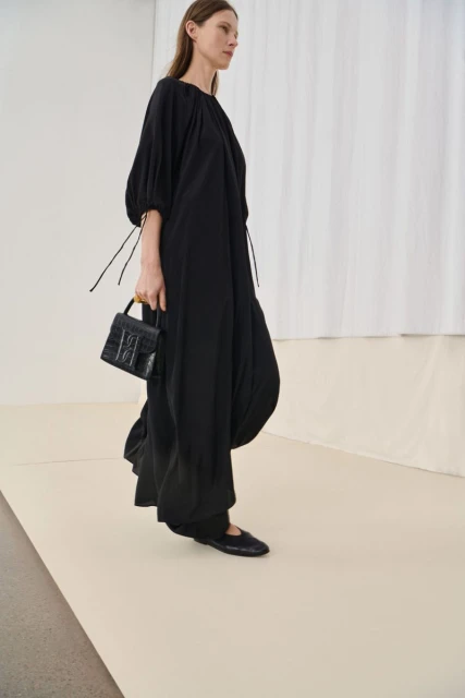 Fashion model in a black dress and matching flats, carrying a leather handbag against a neutral backdrop in a studio environment.