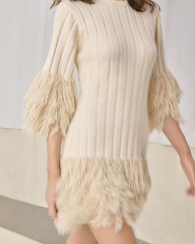 Model wearing a cream ribbed knit mini dress with textured fringe cuffs and hem walking in a minimalist studio setting.