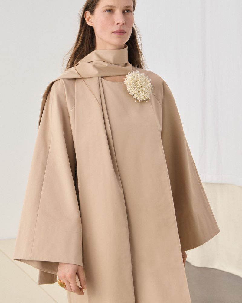 Fashionable adult female poses in an indoor studio wearing a tan dress with flower brooch and shawl, creating an elegant and stylish aesthetic.