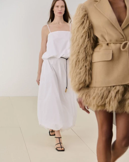 Two women on a runway, one in a white dress and strappy sandals, the other wearing a tan jacket with fur details and light-colored skirt.