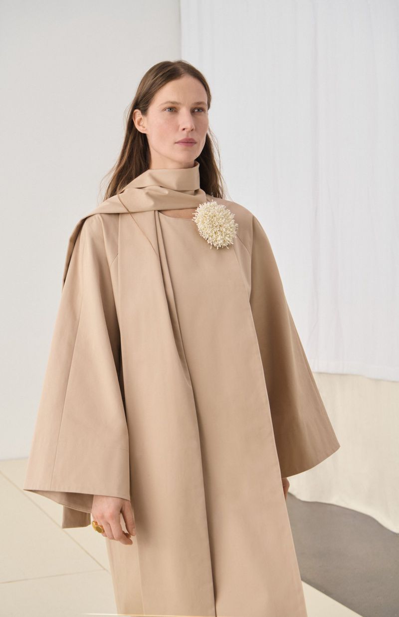 An adult female poses indoors wearing a beige cape dress with a decorative brooch against a blurred white background in a studio setting.