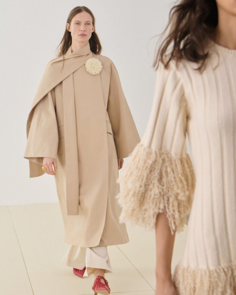 Runway fashion show with a female model wearing a beige coat and white pants, walking on a light yellow floor in a minimalist setting.