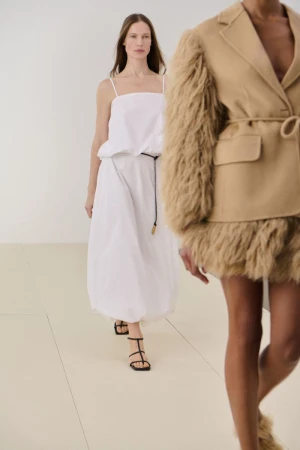 Model walking in a white spaghetti-strap balloon dress alongside a model in a camel jacket with shearling sleeves.