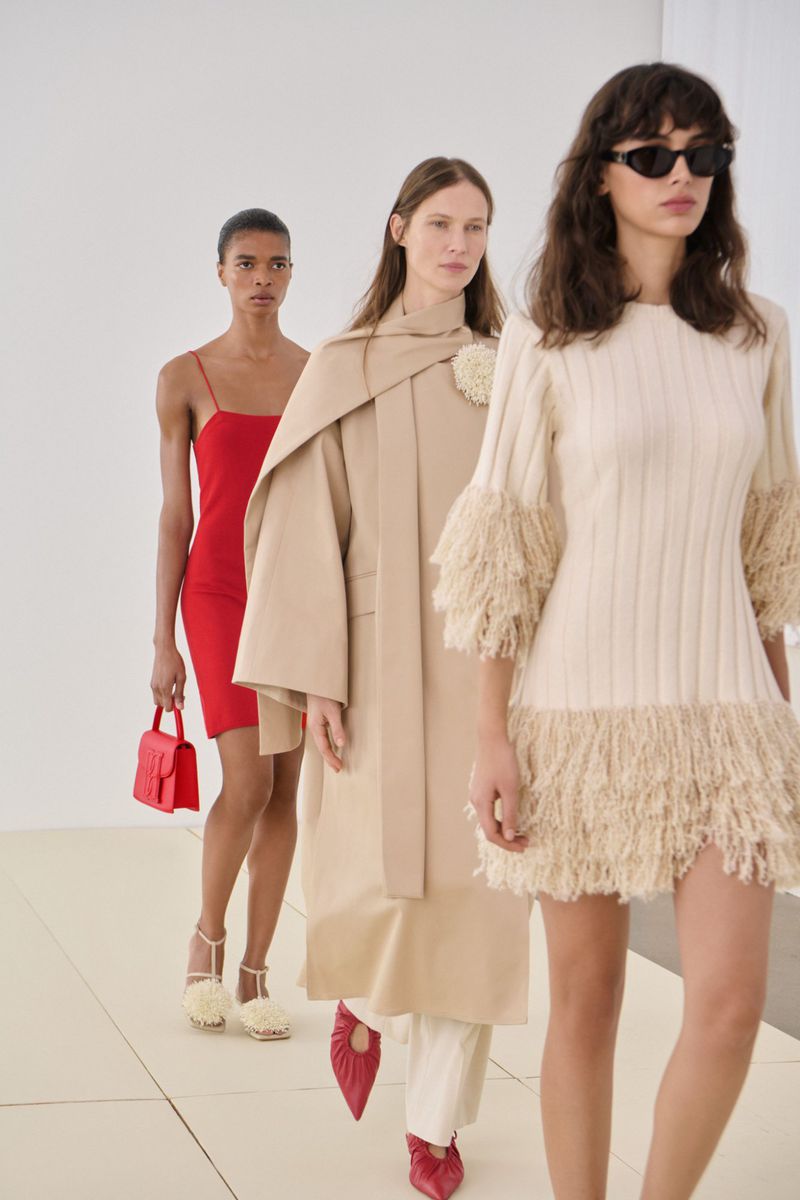 Three fashion models walk on a white runway, wearing red and beige dresses and matching heels, during a runway show.