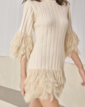 Model wearing a cream ribbed knit mini dress with textured fringe cuffs and hem walking in a minimalist studio setting.